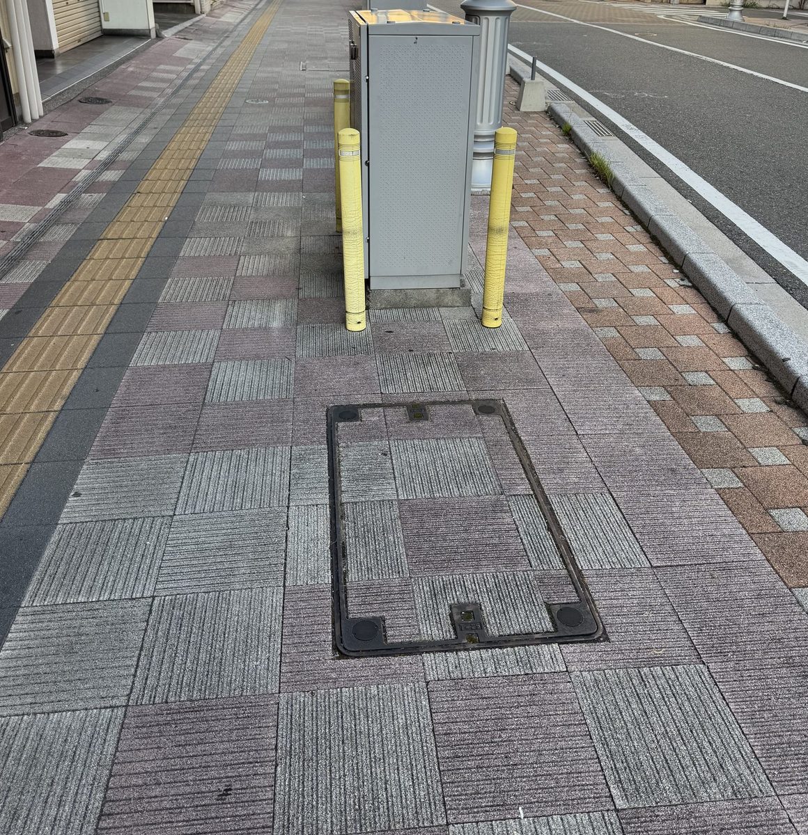 タイル敷きの歩道に、角の丸い長方形の鉄枠がはめ込まれている。枠の内側はまわりと同じ舗装材で埋められていて、へりの一か所に「T-25」の文字が鋳出されている。奥には明るい灰色の四角い地上機器が立ち、その両側に黄色いポールが立っている