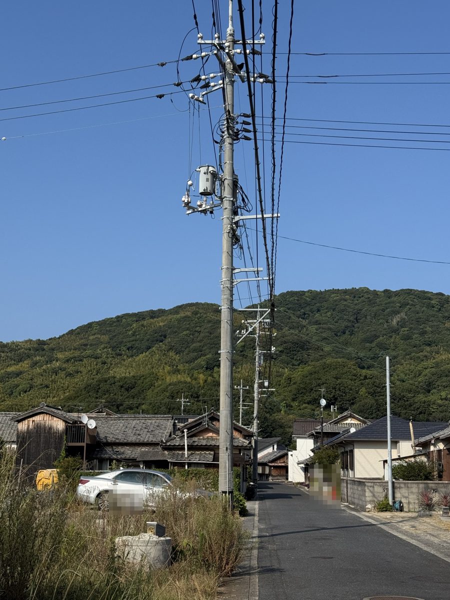 住宅街の通りに立つ電柱の写真。柱の中ほどに灰色の円筒形の柱上変圧器が付き、電線が通り沿いの家々の方向へ伸びている