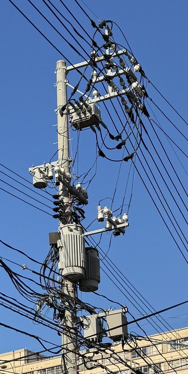 青空を背にした電柱を見上げた写真。いちばん上の腕金には白いがいしがいくつも並び、その下に3つのブッシングが突き出した灰色の箱型の機器、さらに下に白い筒形の部品が集まった金具、その下に縦のひだが入った灰色の円筒形の機器と黒い箱がならぶ。いちばん下では通信のケーブルが何本も束になって横へ走り、灰色の箱もぶら下がっている